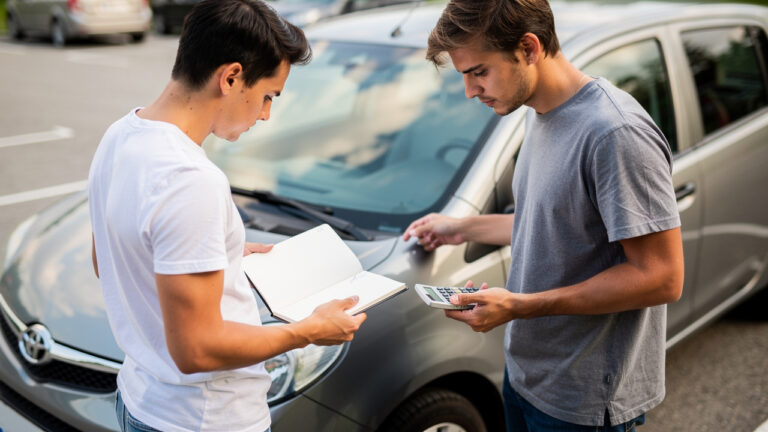 Jeune conducteur examinant attentivement une citadine d’occasion sur un parking, avec un carnet d’entretien ouvert et une calculatrice en main.